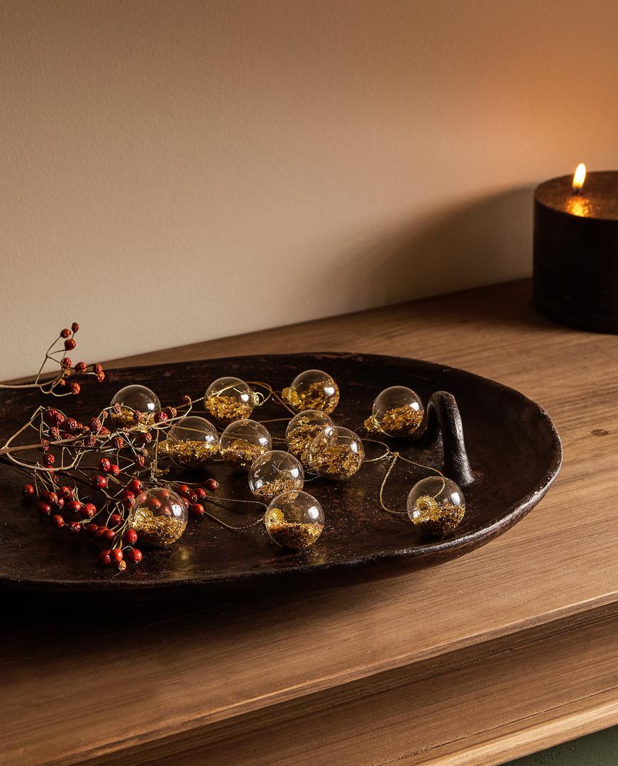 Božićne kuglice punjene zlatnim šljokicama uvučene na vijenac možete objesiti na bor ili ih pak izložiti kao stolnu dekoraciju. Zara Home, 99 kn